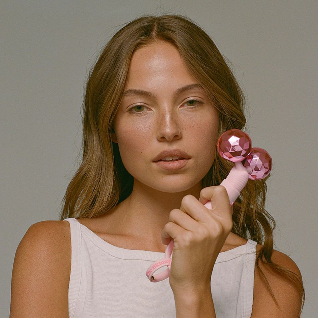 Woman in a white tank top holding a pink facial massage roller with two shiny pink spheres Woman in a white tank top holding a pink facial massage roller with two shiny pink spheres